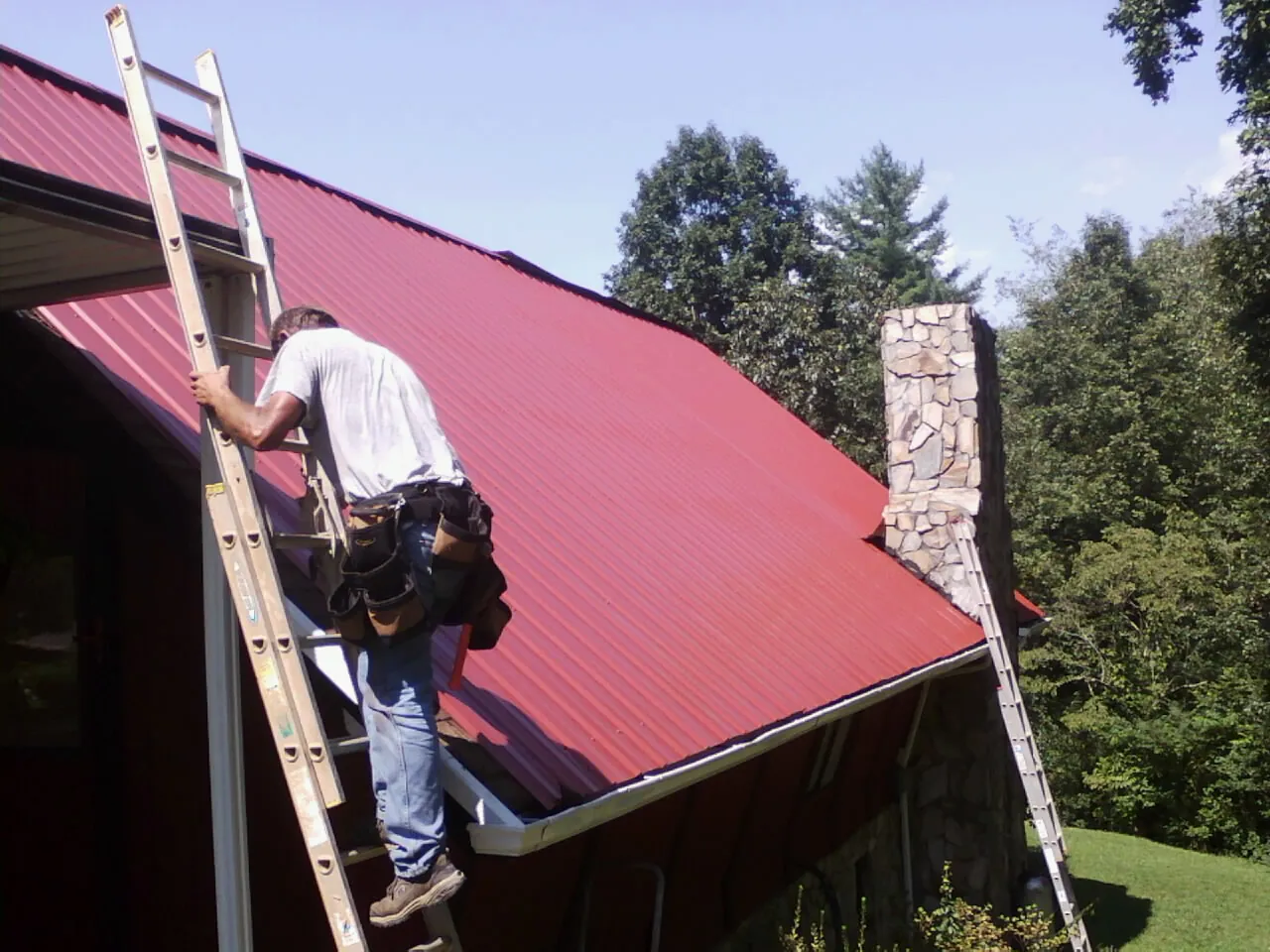 Roofer on a ladder beside a red metal roof and stone chimney — Soffit & Fascia Repair by Roofing Brothers in Snoqualmie Pass